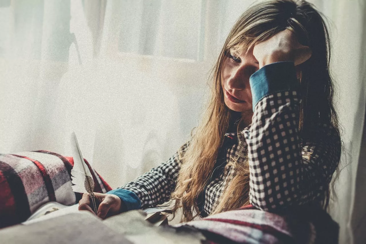 A thoughtful woman sits at a desk, holding a pen in one hand and resting her head in the other, as she contemplates stress and autoimmune health issues.