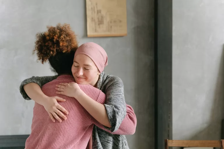 Two women embracing warmly; one wears a pink head scarf, symbolizing support and solidarity, possibly in a context related to health or cancer awareness.